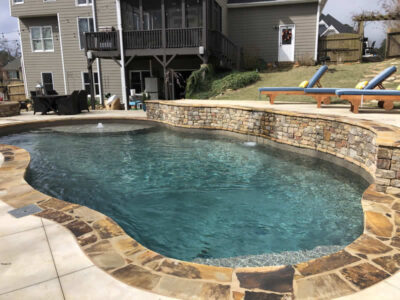 Backyard pool with stone deck and chairs.